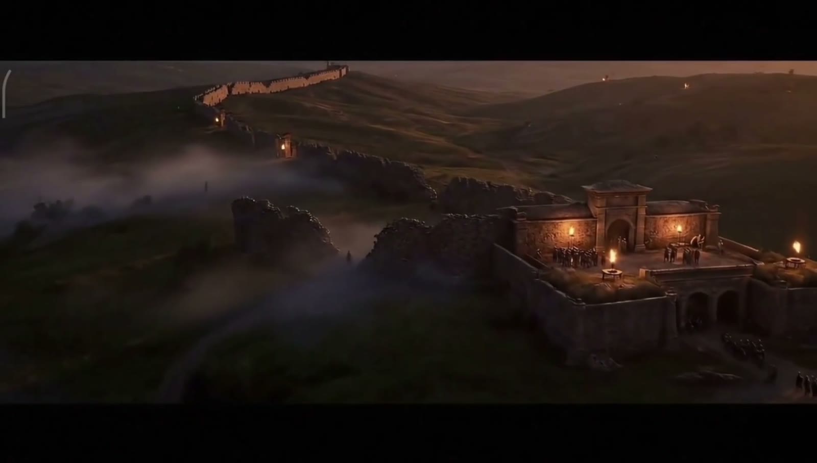 A fortress on a hillside at dusk, torches lit along its walls